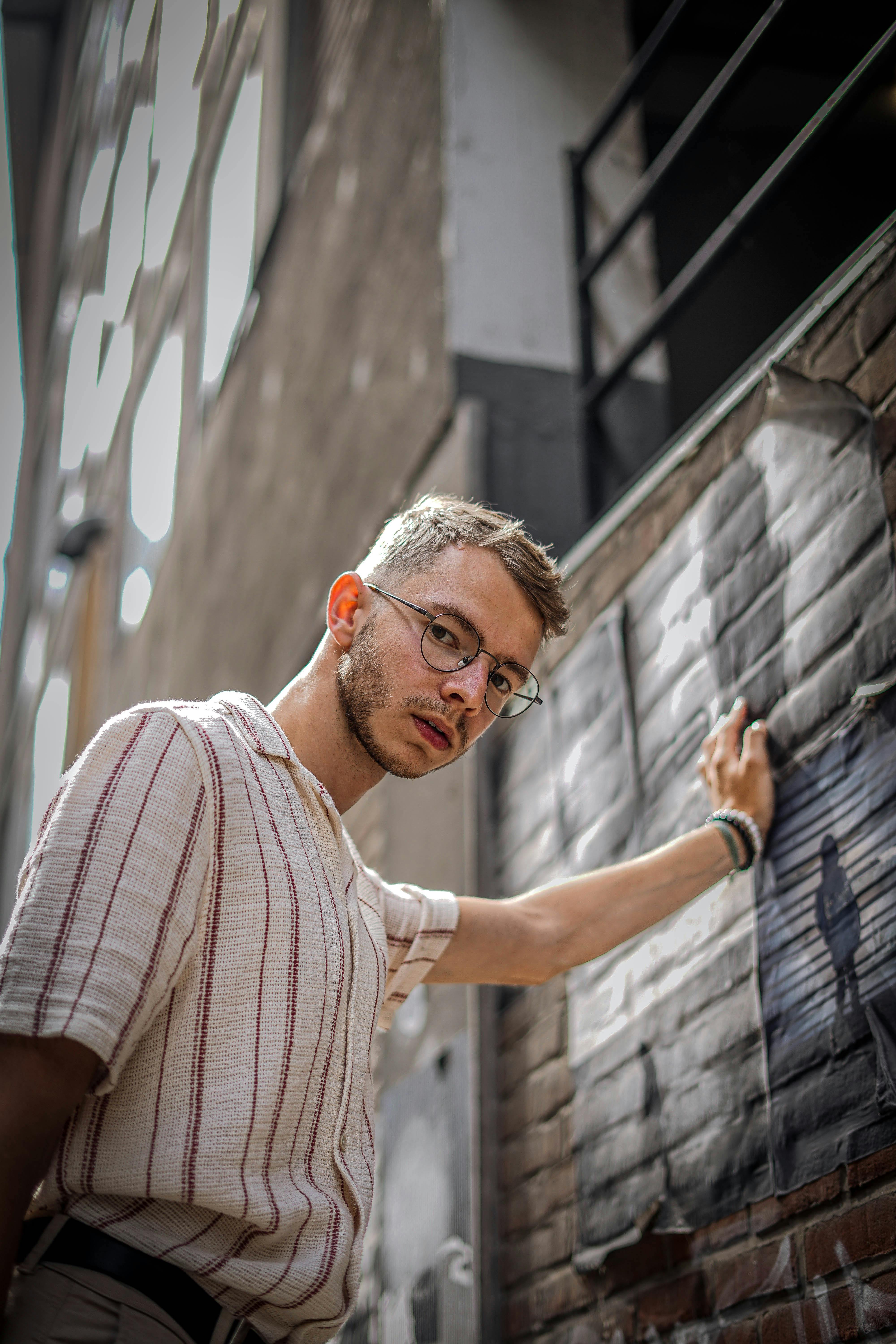 A stylish young man in glasses and a striped shirt poses in an urban alleyway.