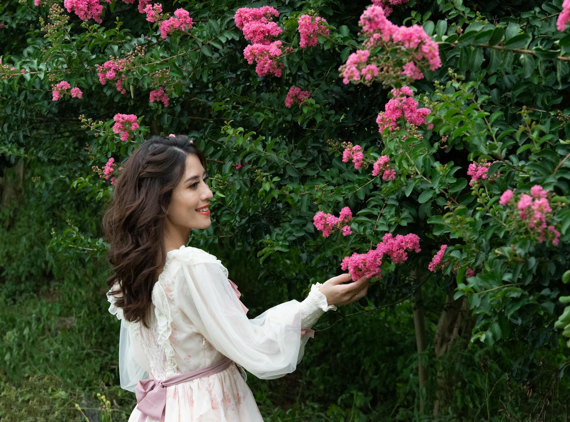 Elegant woman in Edwardian dress admiring crepe myrtle blooms in garden.