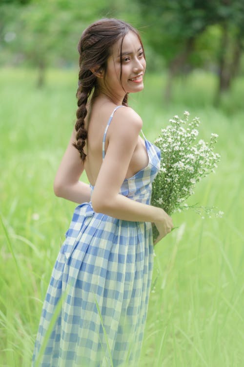 Free Close-Up Shot of a Beautiful Woman in Blue Plaid Dress Holding White Flowers while Standing on Green Grass
 Stock Photo