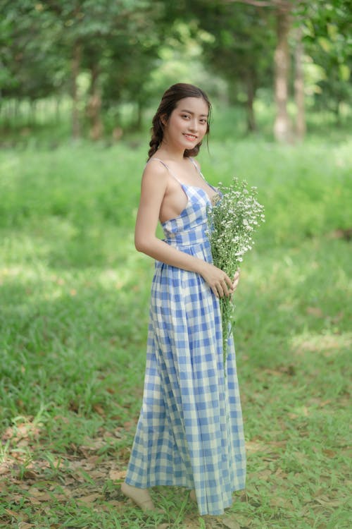 Free Beautiful Woman in Blue Plaid Dress Holding White Flowers while Standing on Green Grass
 Stock Photo