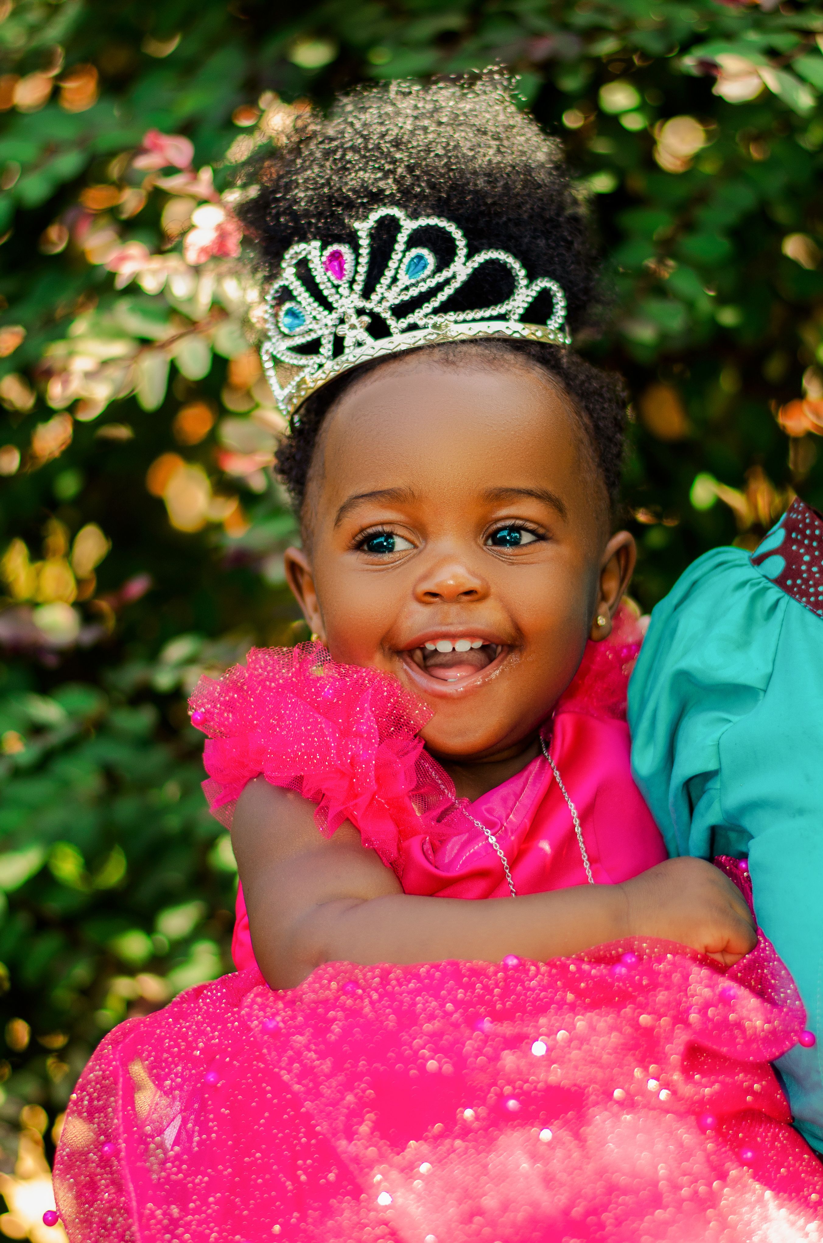 Adorable African American baby girl wearing a pink dress and tiara, smiling outdoors.