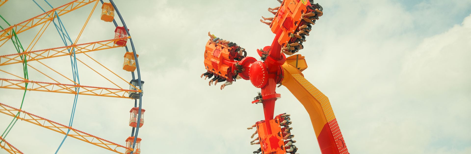 Exciting view of a thrilling amusement park ride next to a colorful Ferris wheel under cloudy skies.