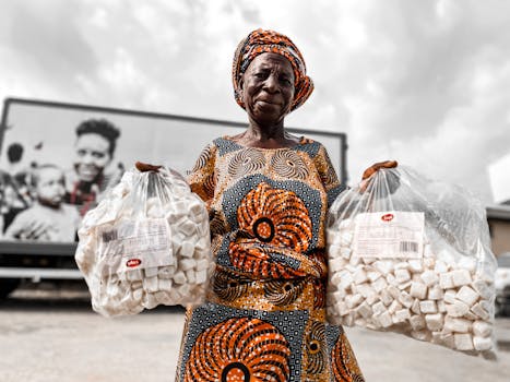 African woman in traditional attire displaying bags of local delicacy at an outdoor market.