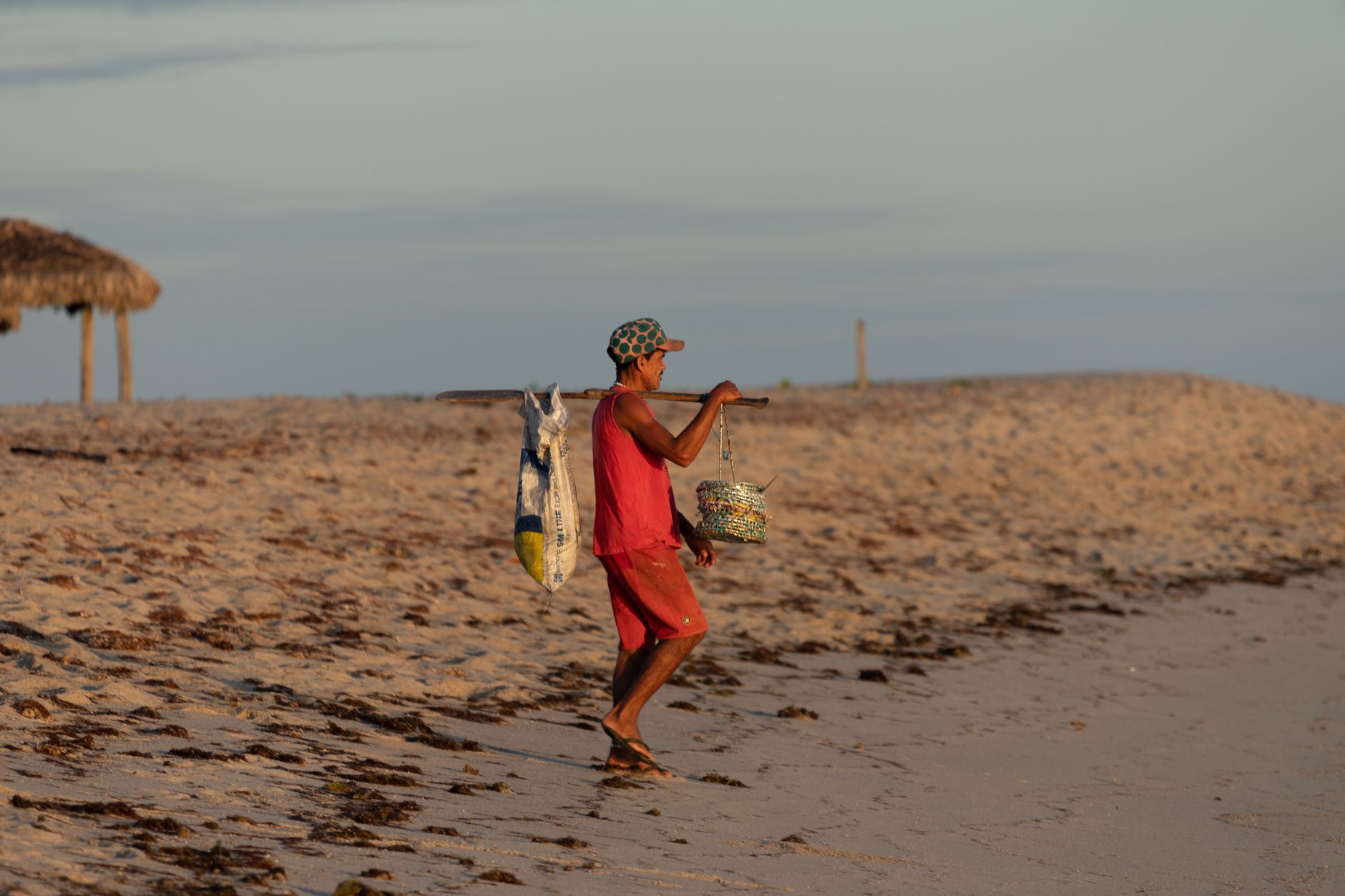 A man in casual clothes walking along a beach holding a pole with bags during sunset.
