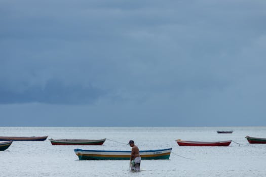 Man stands in water near colorful docked boats on a calm sea under cloudy skies.