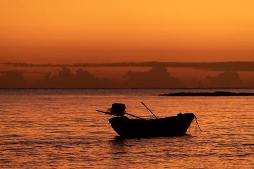 Free A peaceful silhouette of a boat against a vibrant orange sunset over calm sea waters. Stock Photo
