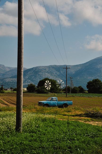 A blue pickup truck parked in a scenic Greek field with mountains in the background.