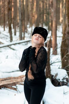 A woman dressed warmly in black, standing in a snow-covered forest, enjoying winter's beauty.