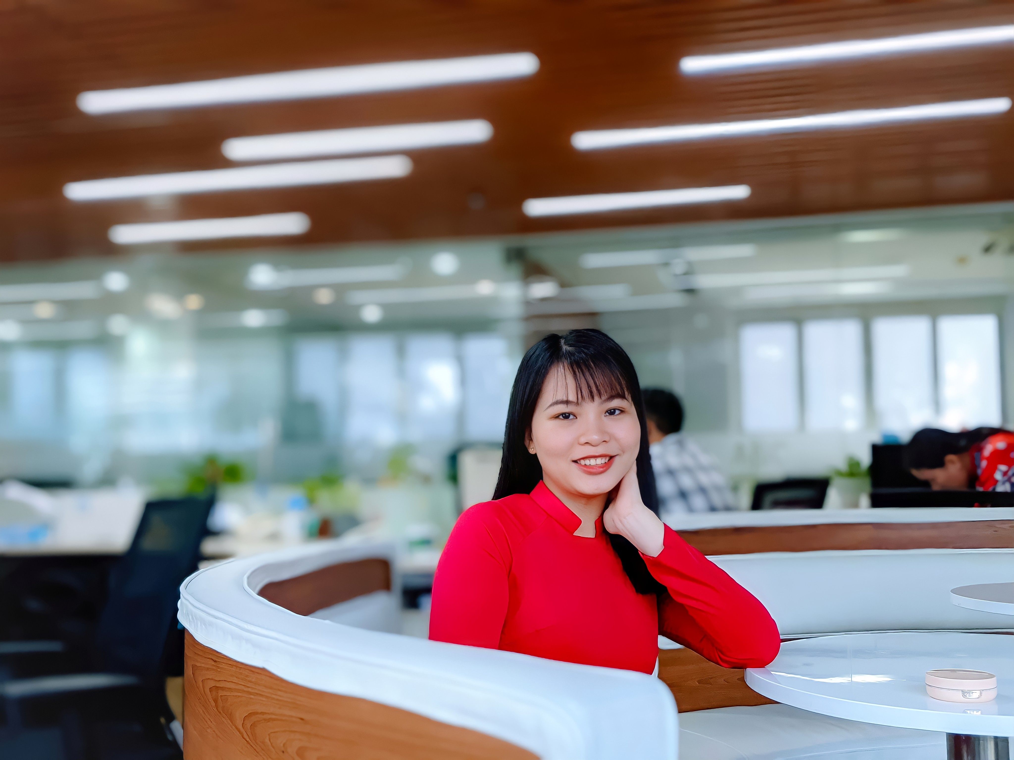 A cheerful woman posing in a modern office, wearing a red blouse and smiling confidently.