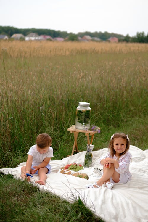 Two Children Sitting on White Picnic Blanket near Grass Field 