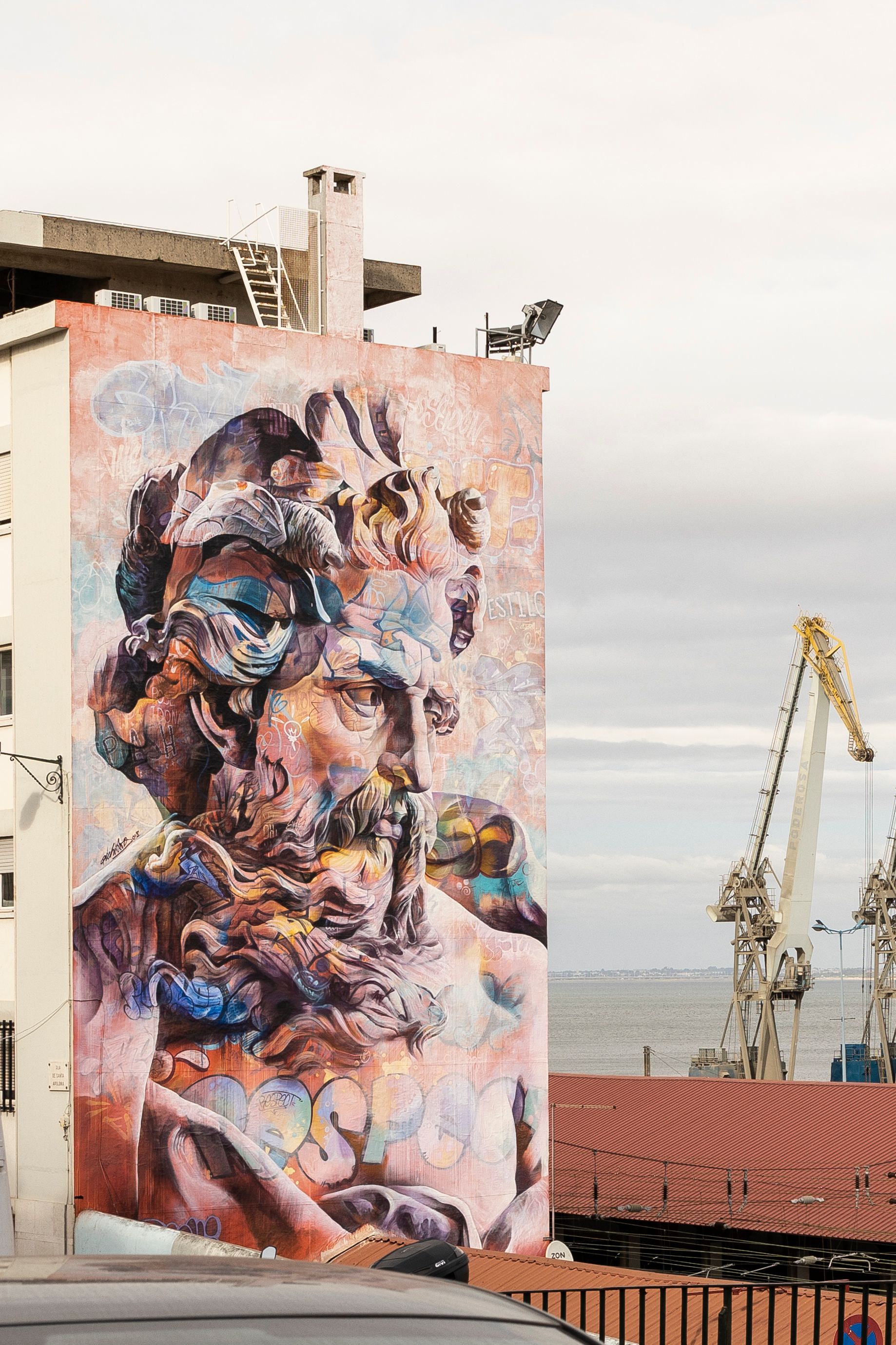 Colorful mural on a residential building by the sea, featuring cranes and urban architecture.