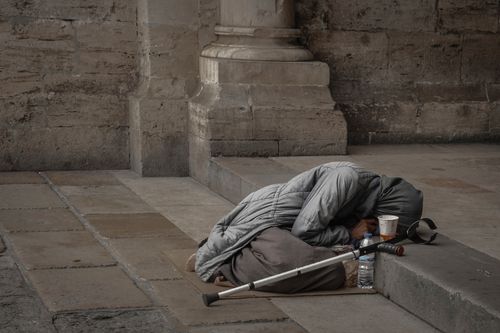 Free A homeless woman in a gray jacket with a hijab rests against a stone wall, highlighting urban poverty. Stock Photo