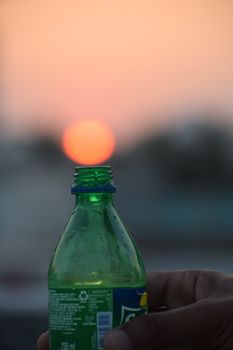 A close-up of a hand holding a bottle with a sunset in the background.
