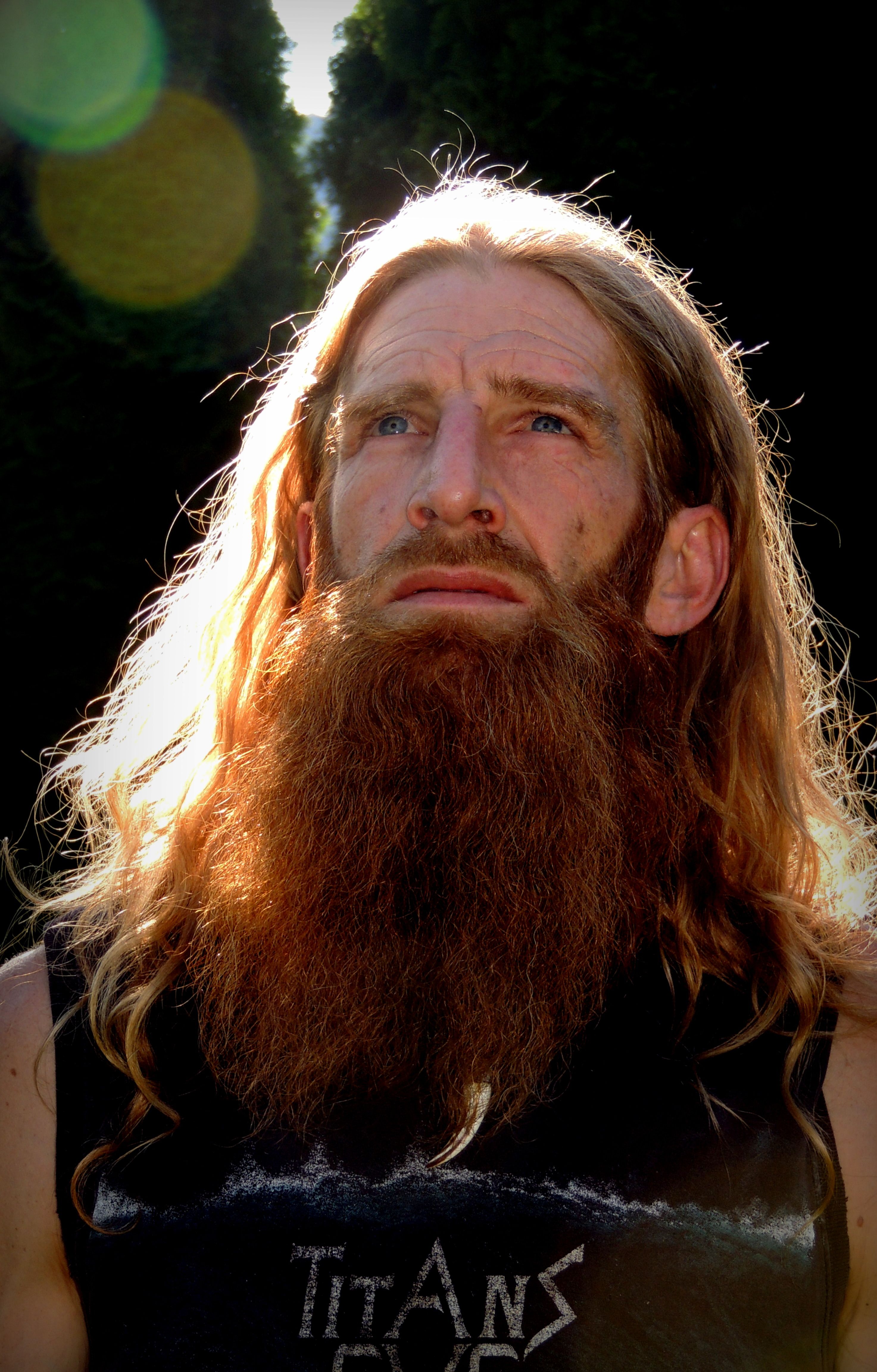 Close-up portrait of a man with long hair and a beard looking up in bright sunlight.