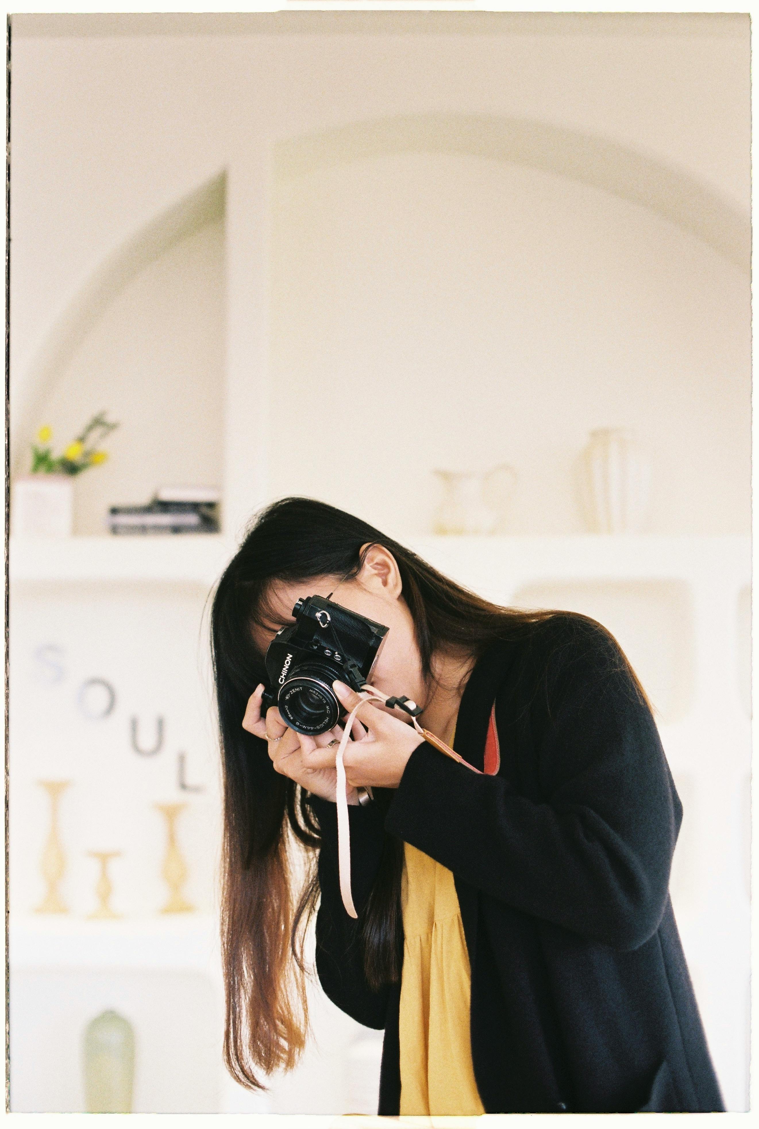 A young woman in black attire holding a camera creatively indoors.