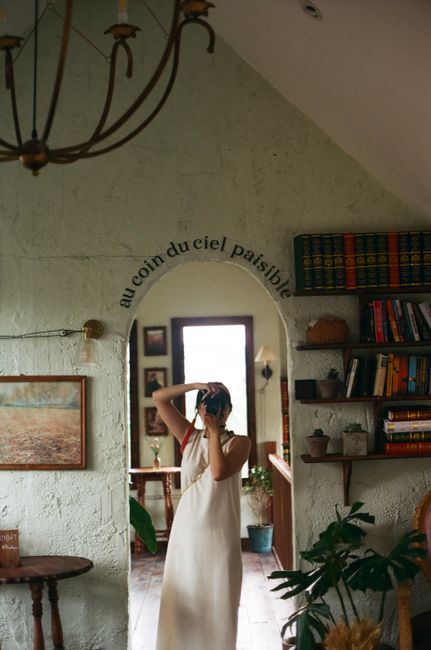 A woman in a white dress taking a picture in a book-filled room, reflecting in a mirror.