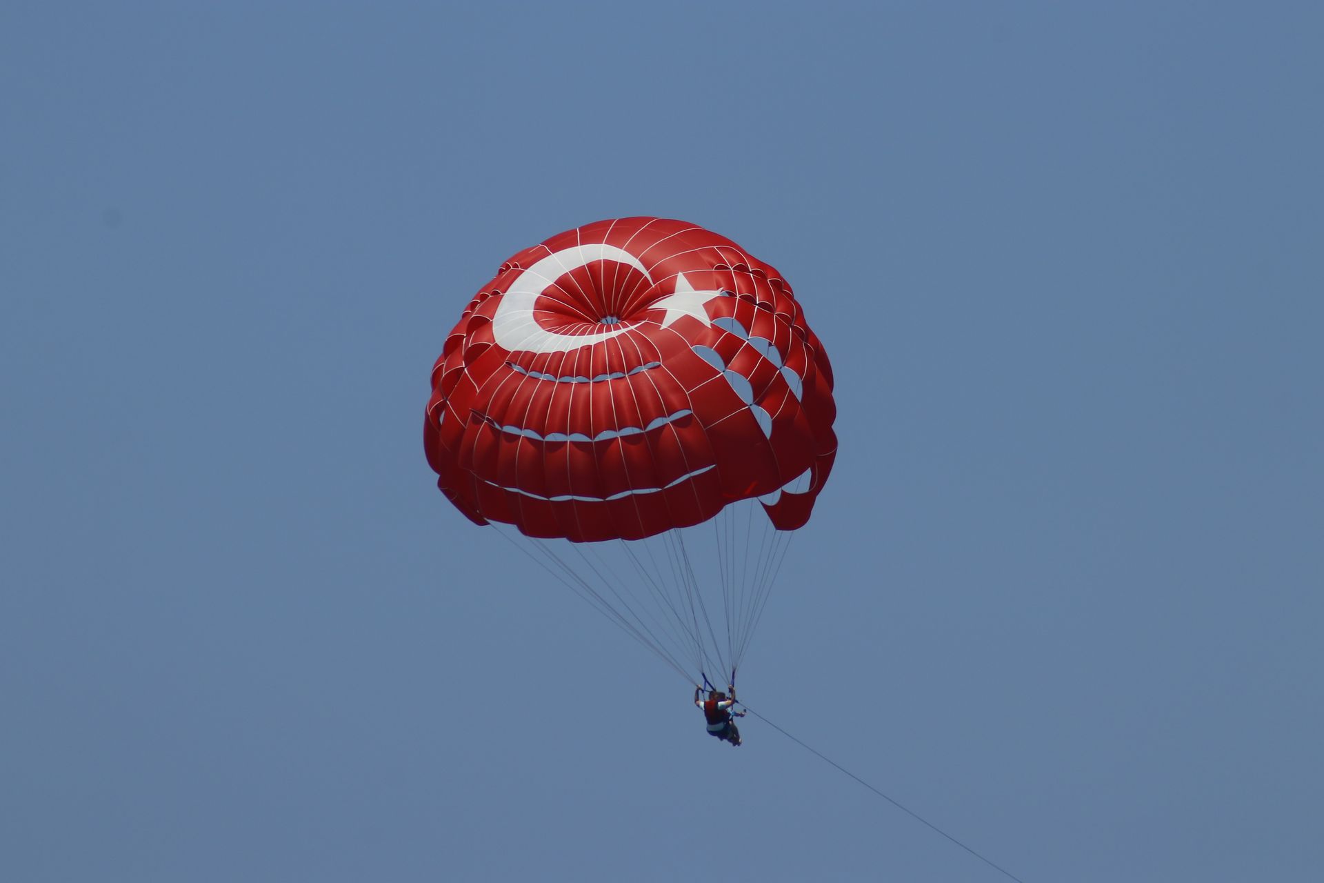 Person parasailing with a red parachute featuring a Turkish flag against a clear blue sky.