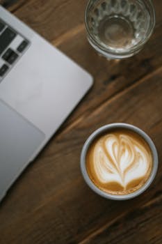 Close-up of a latte with heart-shaped latte art on a rustic wooden table alongside a laptop.