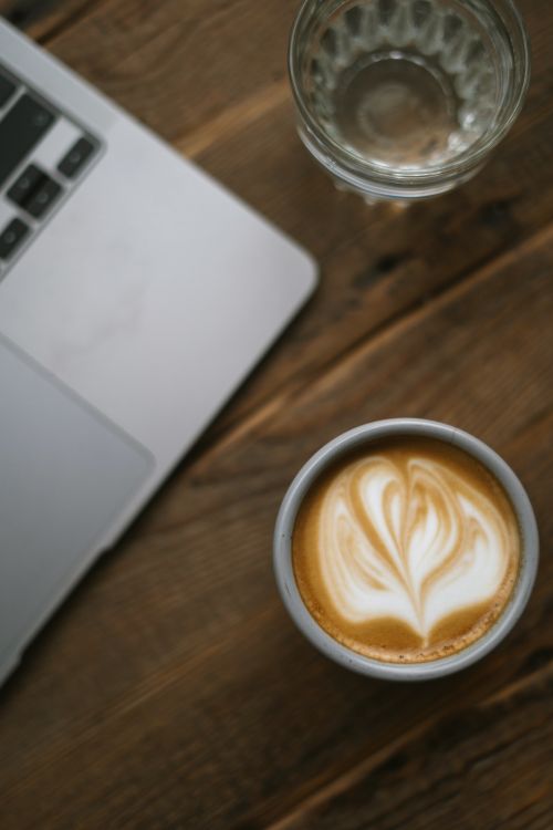 Free Close-up of a latte with heart-shaped latte art on a rustic wooden table alongside a laptop. Stock Photo