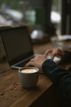 Person working on a laptop in a café with a cappuccino on a wooden table.