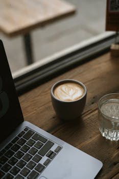 A cozy café scene with a laptop and latte on a wooden table by the window.