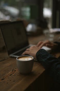 Hands typing on a laptop beside a steaming cup of coffee on a wooden table.