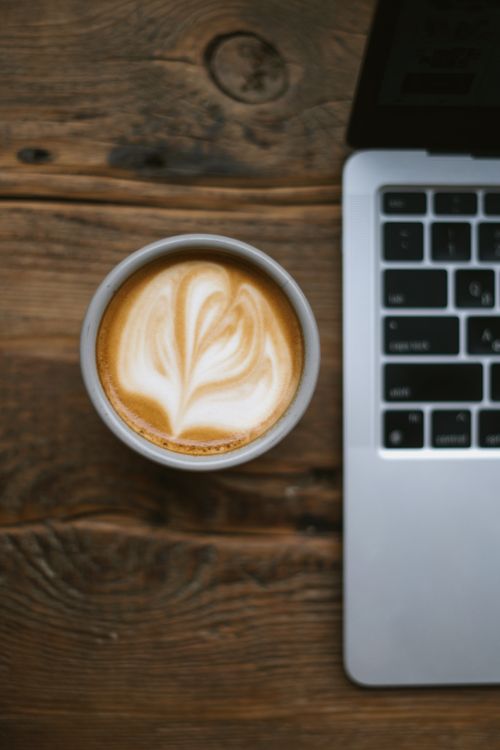 Free A latte with art next to a laptop on a wooden table, perfect for workspace themes. Stock Photo