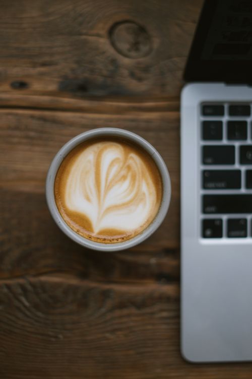 Free Stylish latte art in a cup next to a laptop on a rustic wooden table. Perfect workspace vibe. Stock Photo