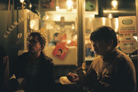 Two men enjoy takeout food at night outside a street food spot in Athens, Greece.