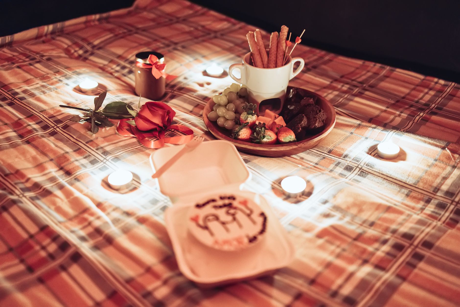 Cozy candlelit picnic setup with delicious desserts, fruits, and a red rose on plaid blanket.