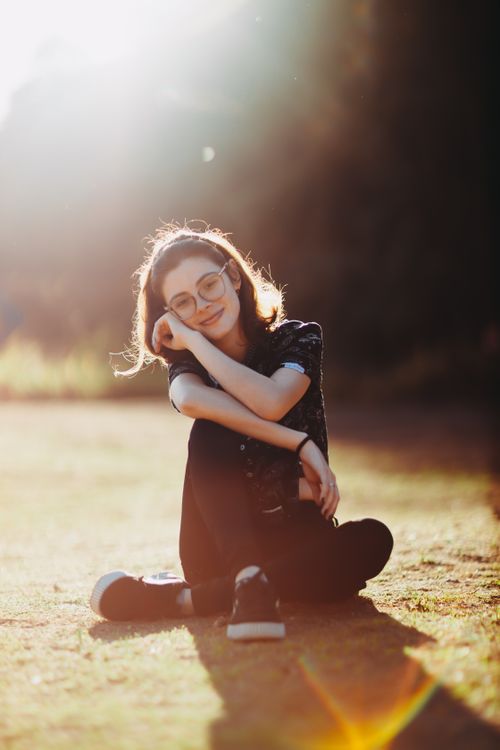 Free Cheerful young woman with eyeglasses sitting on grass, enjoying the sunlight. Stock Photo