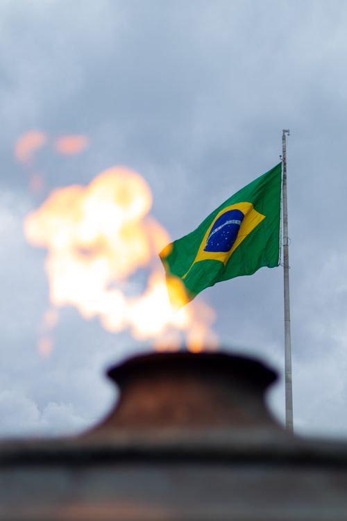 Free The Brazilian flag waves atop a flagpole with a fiery flame in the foreground against a cloudy sky backdrop. Stock Photo