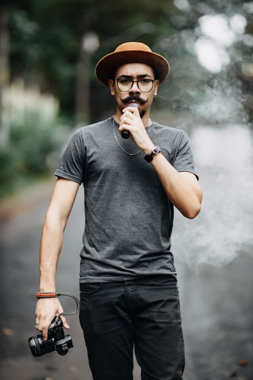 Free Fashionable man with mustache, brown hat, and camera vaping on a street. Stock Photo