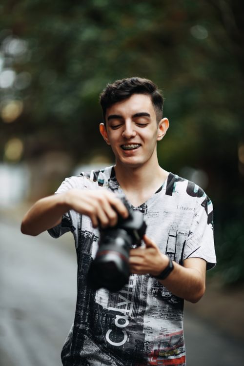 Free A young man smiling while holding a DSLR camera outdoors, creating a joyful mood. Stock Photo