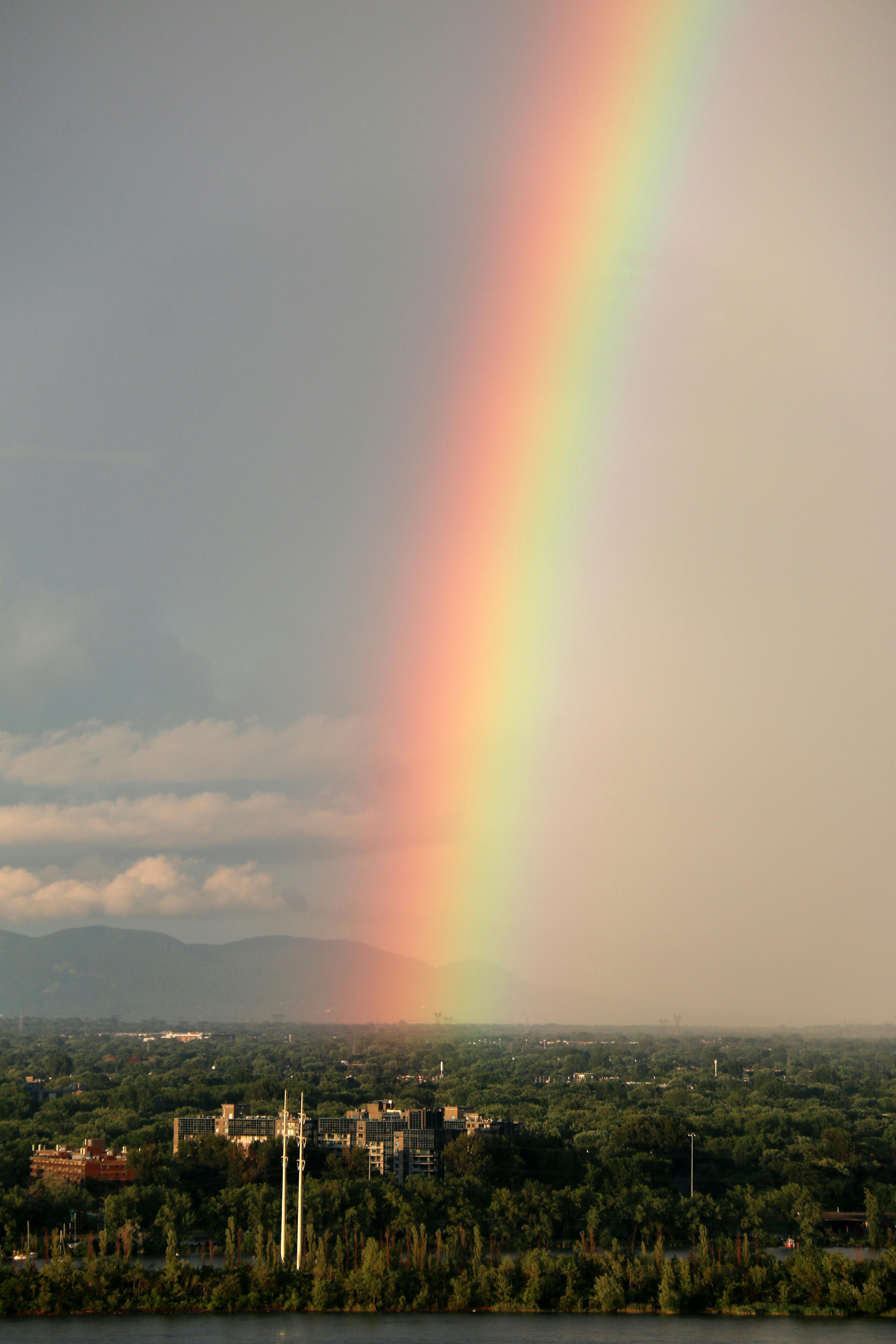 A stunning rainbow arches over buildings with trees and mountains in the background at dusk.