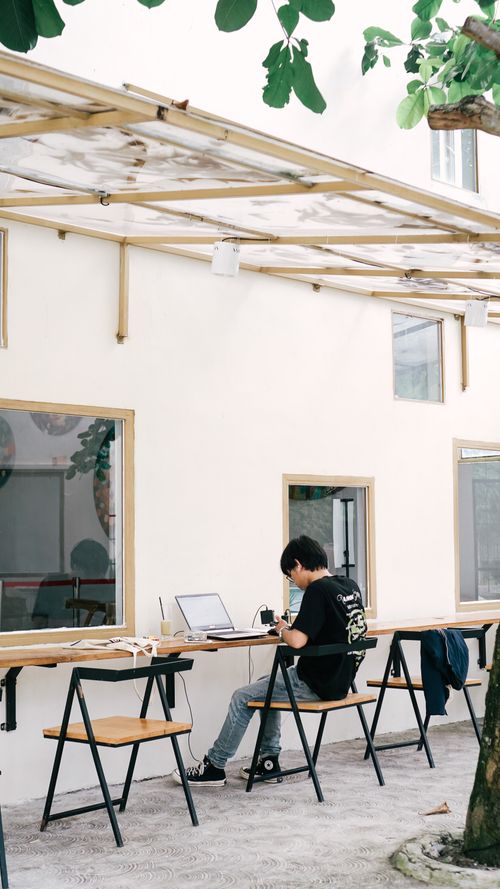Free A young person working on a laptop in an outdoor cafe setting under a canopy surrounded by trees. Stock Photo