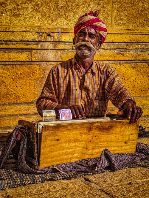 Free Portrait of an elderly Indian man with a mustache, wearing a turban and sitting on a sidewalk with a wooden box and money in front of a yellow background. Stock Photo