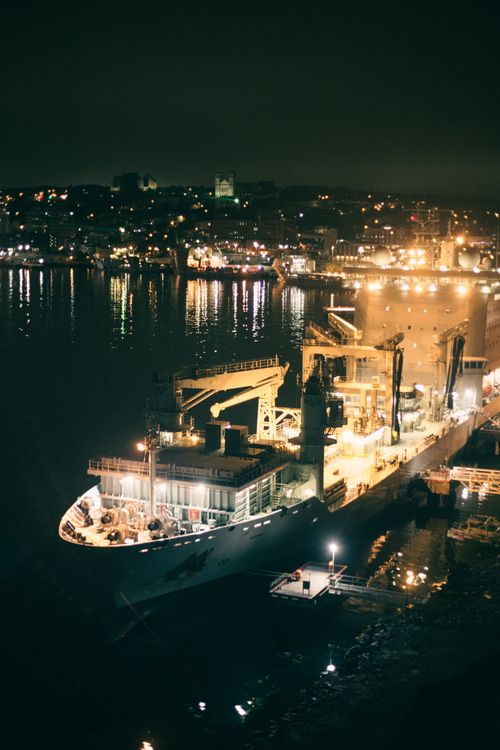 Free A brightly lit cargo ship docked at a busy harbor with city lights reflecting on the water. Stock Photo