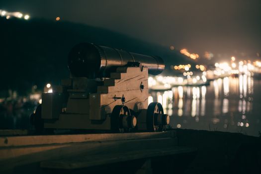 A vintage cannon positioned by the water with city lights reflecting in the harbor at night.