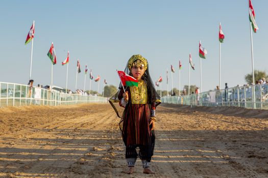 A young girl in traditional Omani clothing holds the flag of Oman during a festive outdoor celebration.