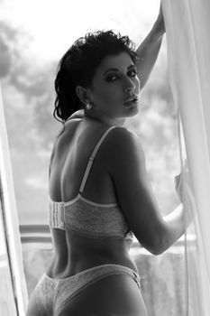 Black-and-white portrait of a woman posing in elegant lingerie by a window.