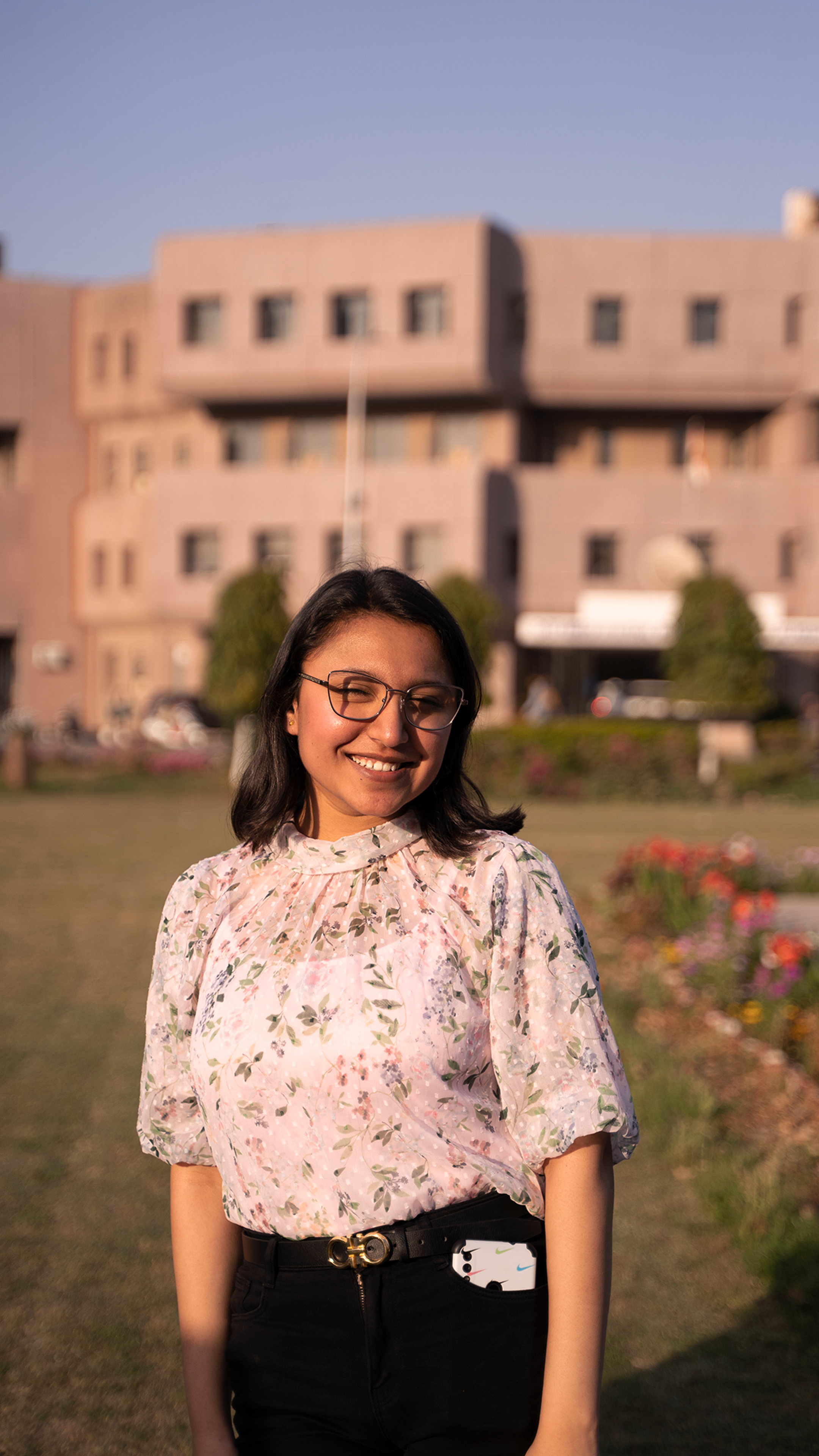 A young woman with eyeglasses smiling outdoors, with a blurred building backdrop.