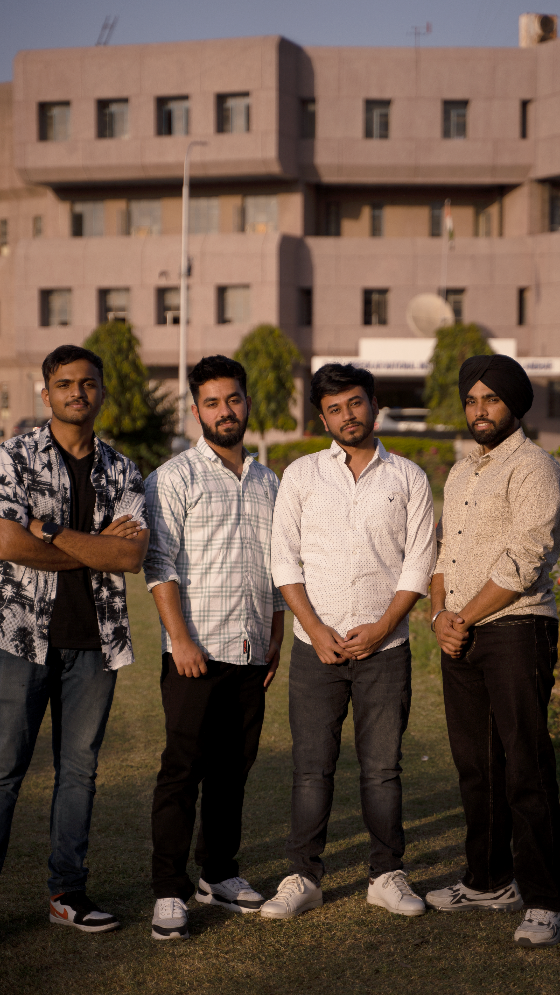 Four friends in casual attire stand on grass in front of a building, smiling.