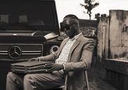 Man in sunglasses sitting outdoors with suitcase near Mercedes-Benz car.