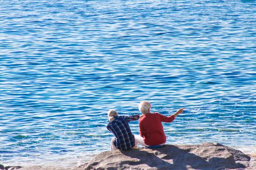 Free Two individuals enjoying a peaceful moment sitting on rocks by a tranquil blue sea. Stock Photo