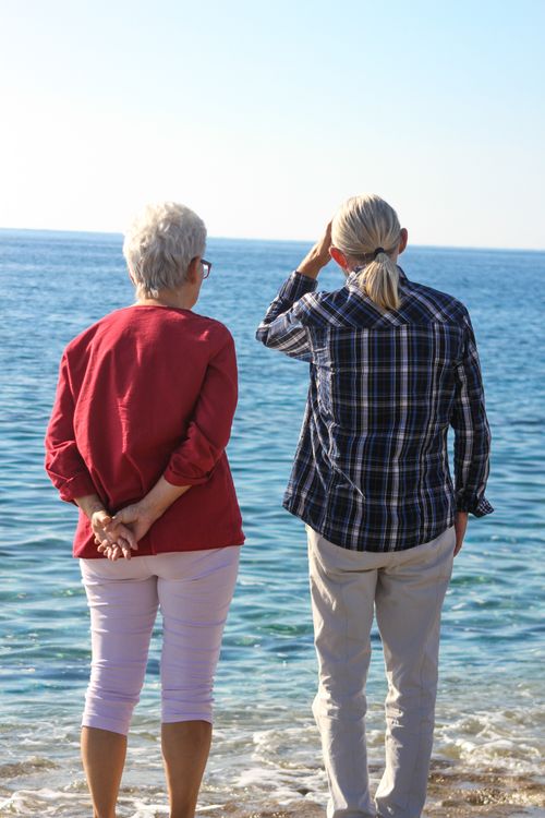 Free Two senior women stand on a serene beach, gazing at the calm sea under the clear blue sky. Stock Photo