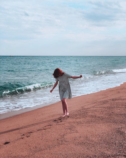 Woman Wearing a Striped Dress Walking Barefoot on Shore