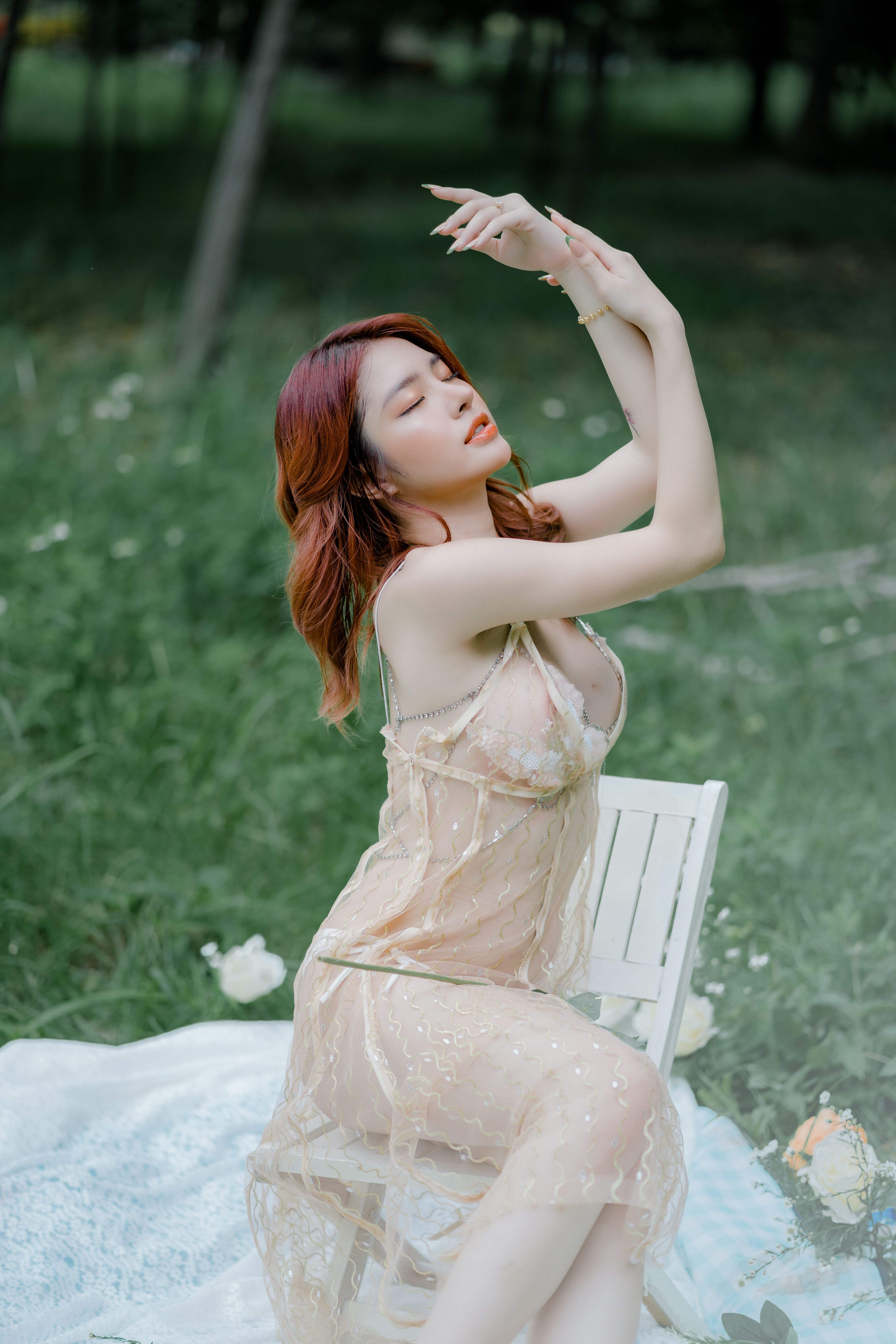 Asian woman in a sheer dress posing elegantly on a chair in a grassy field.