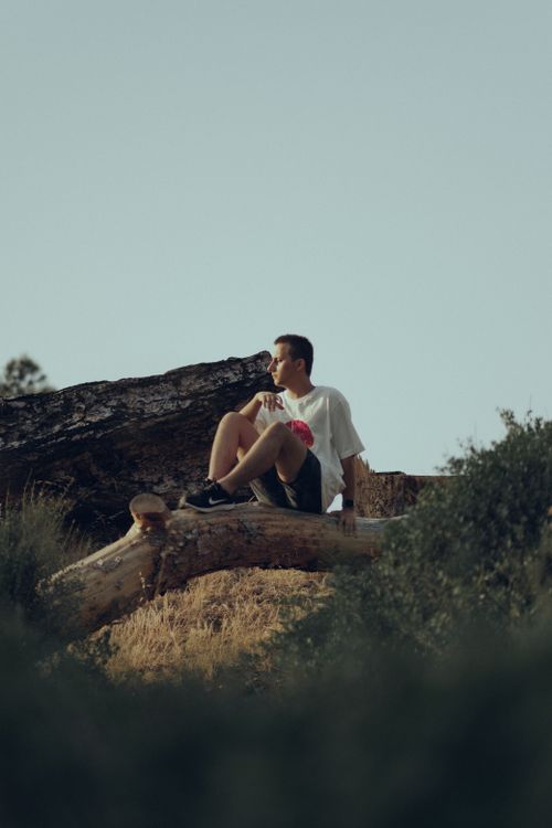 Free A man in a white t-shirt sits on a log outdoors, enjoying a peaceful moment in nature. Stock Photo