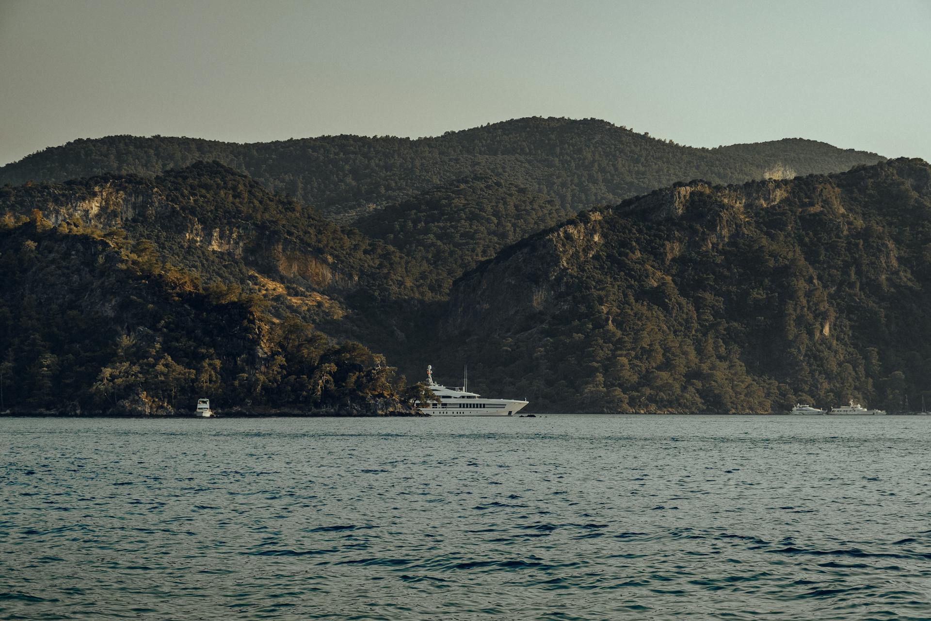 A luxury yacht sails along a rugged, scenic coastline under a calm sky.
